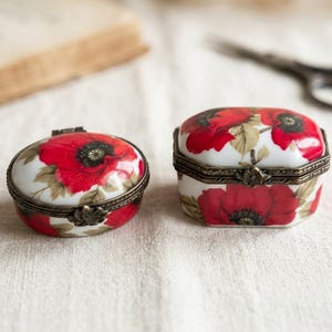 May include: Two small porcelain trinket boxes with a white base and a red poppy flower pattern. Each box features a hinged lid with an antique brass-toned metal rim and a decorative clasp. One box is oval-shaped, and the other is octagonal.