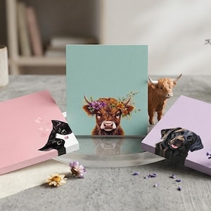 Könnte beinhalten: Drei quadratische Haftnotizblöcke mit Tierillustrationen. Ein rosa Block zeigt eine schwarz-weiße Katze, ein hellblauer Block in der Mitte zeigt ein Hochlandrind mit Blumenkranz und ein lavendelfarbener Block zeigt einen fröhlichen schwarzen Hund.
