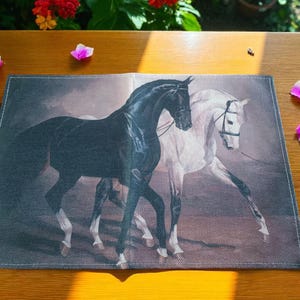 Peut inclure: Nappe rectangulaire avec une peinture de chevaux noir et blanc. Les chevaux sont représentés côte à côte. La nappe est posée sur une table en bois, avec des pétales de fleurs roses éparpillés.
