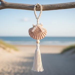 Può includere: Un ornamento a forma di conchiglia sospesa con una nappa color crema e perline di legno. La conchiglia è di colore pesca chiaro, con un nastro bianco e dettagli in pizzo. L'ornamento è appeso a un ramo di legno, con una spiaggia e un oceano sfocati sullo sfondo.