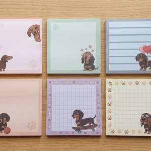 Puede incluir: Seis blocs de notas adhesivas cuadrados dispuestos en dos filas. Cada bloc presenta un color pastel diferente y una ilustración única de un perro salchicha. Los diseños incluyen un perro con una pelota, en un monopatín, con globos y bordes de huellas.