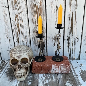 May include: A decorative skull with etched details sits next to two black metal candle holders with yellow candles. The candle holders are adorned with vintage keys and sit on a red brick. The background is a weathered white wooden surface.