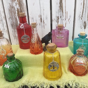 Peut inclure: Plusieurs bouteilles décoratives en verre de différentes couleurs, notamment orange, rouge, vert, jaune, rose et bleu. Chaque bouteille est ornée d'un bouchon en liège et d'un charm argenté. Elles sont disposées sur un tissu jaune.