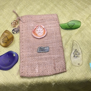 May include: A burlap pouch with a shell charm and a label that reads "Four Elements". Four glass stones with symbols representing the elements of fire, earth, air, and water are arranged around the pouch.