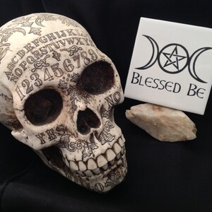 May include: A white skull with a black Ouija board design on the front. The skull is sitting on a black surface. A white tile with a black triple moon symbol and the text "Blessed Be" is sitting on a white rock.