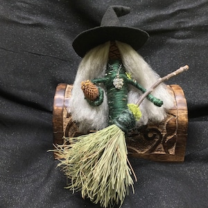 May include: A green witch doll with long white hair, a black pointed hat, and a broom. The doll is sitting on a wooden box with carved details. The doll is holding a small pine cone in one hand and a twig in the other.