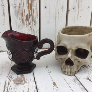 Op de afbeelding: Een donkerrode glazen roomkan met een zilveren pentagram-ontwerp en een witte keramische schedel-plantenpot.