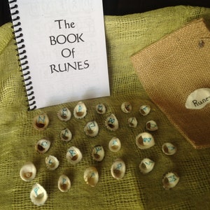 May include: A set of 24 runes made from seashells with blue painted symbols on a green fabric background. The runes are in a burlap bag with the word "Runes" written on it. A book titled "The Book of Runes" is also in the image.