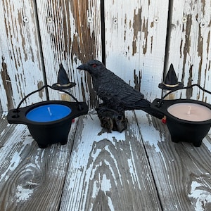 Puede incluir: Velas negras en forma de caldero con cera azul y beige, con una estatua de cuervo negro con ojos rojos. Sombreros de bruja en miniatura sobre botellas de vidrio, creando una exhibición espeluznante con temática de Halloween.