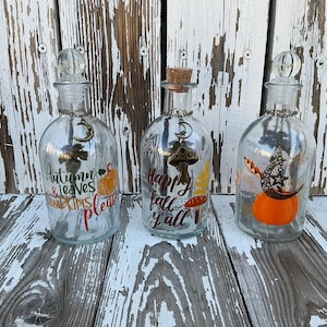 May include: Three clear glass bottles with decorative charms and stoppers. Each bottle has autumn-themed text and imagery, including pumpkins, leaves, and a mushroom. The bottles are displayed on a weathered white wooden surface.