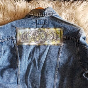 Op de afbeelding: Een blauwe denimjas met een kleurrijke patch op de rug. De patch heeft een cirkelvormig ontwerp met een mandala-achtig patroon in tinten groen, geel en bruin.