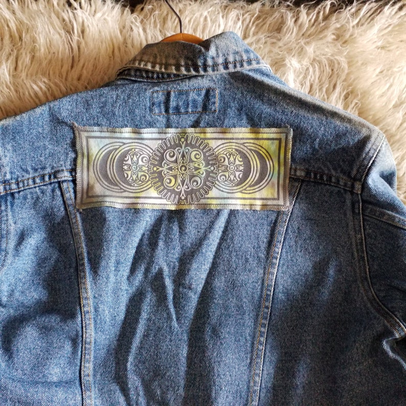 Op de afbeelding: Een blauwe denimjas met een kleurrijke patch op de rug. De patch heeft een cirkelvormig ontwerp met een mandala-achtig patroon in tinten groen, geel en bruin.