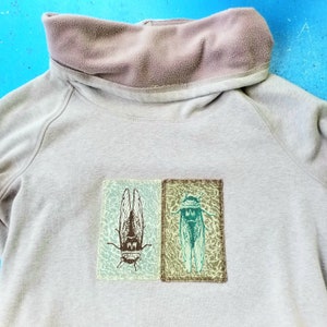 Peut inclure: Un sweat-shirt gris clair avec un col en polaire marron et deux patchs brodés de cigales sur le devant. Les cigales sont vertes et brunes avec des ailes détaillées.