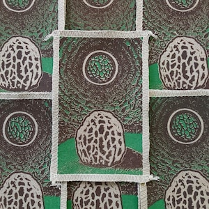 May include: A repeating pattern of fabric squares featuring a morel mushroom illustration in brown and white. Each square has a green border and a central design of a green circle. The background is a textured dark brown.