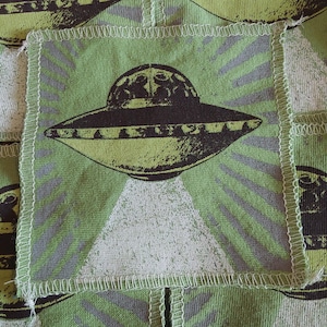 Pode incluir: Tecido verde com um padrão repetido de OVNIs. Cada OVNI é castanho e verde limão, com um feixe de luz branco. O fundo é verde claro com raios cinzentos.