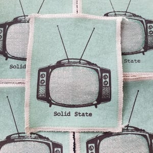 Könnte beinhalten: Ein Stapel hellblauer Stoffpatches mit einer schwarzen und weißen Vintage-Fernsehdarstellung und dem Text "Solid State".