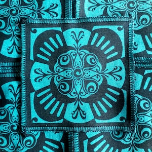 May include: A close-up of a teal and black fabric with a repeating pattern of ornate floral designs. The fabric is stitched together in a patchwork style.