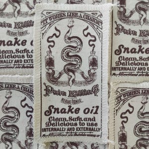 Pode incluir: Um emblema retangular de tecido com fundo creme e tinta castanho-escura. O design apresenta uma cobra central enrolada em torno de duas garrafas, com o texto: IT WORKS LIKE A CHARM, Pain Killing NERVE TONIC, Snake oil, Clean, Safe, and Delicious to use INTERNALLY AND EXTERNALLY.