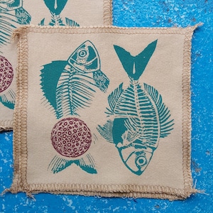 May include: Square beige fabric coasters with teal fish skeleton designs and a maroon circular pattern. The coasters have a stitched border and are arranged on a blue surface. The design is a unique, artistic representation of marine life.