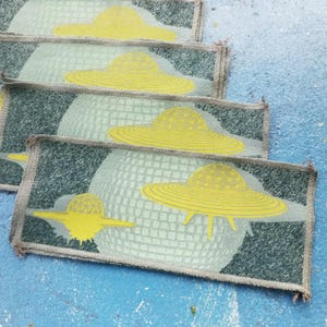 May include: A stack of rectangular fabric patches with a retro design. Each patch features a yellow flying saucer over a light green sphere against a dark green background. The patches have a textured appearance.