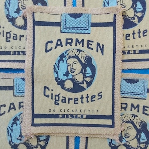 May include: Tan fabric patch with a vintage design featuring the text "CARMEN Cigarettes" and an illustration of a woman's face in a blue circle. The patch has a blue border and a beige fringe.