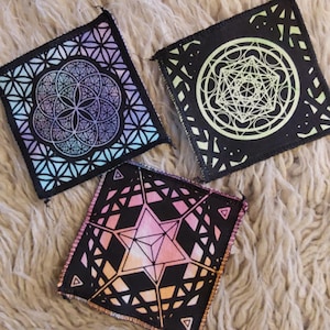 Può includere: Tre toppe quadrate con disegni geometrici. Una presenta un motivo a fiore della vita in blu, viola e verde acqua. Un'altra ha un disegno del Cubo di Metatron in bianco e verde. La terza toppa mostra un disegno a stella in rosa e arancione.