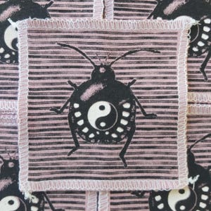 May include: Square patch with a black bug design and a yin-yang symbol on a pink background with black horizontal stripes. The patch is bordered with white stitching. The design is repeated across the fabric.