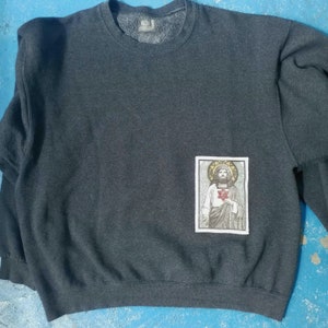 May include: Dark grey sweatshirt with a rectangular patch showing a religious illustration. The illustration depicts a figure with a halo, holding a heart. The sweatshirt has a crew neck and long sleeves.