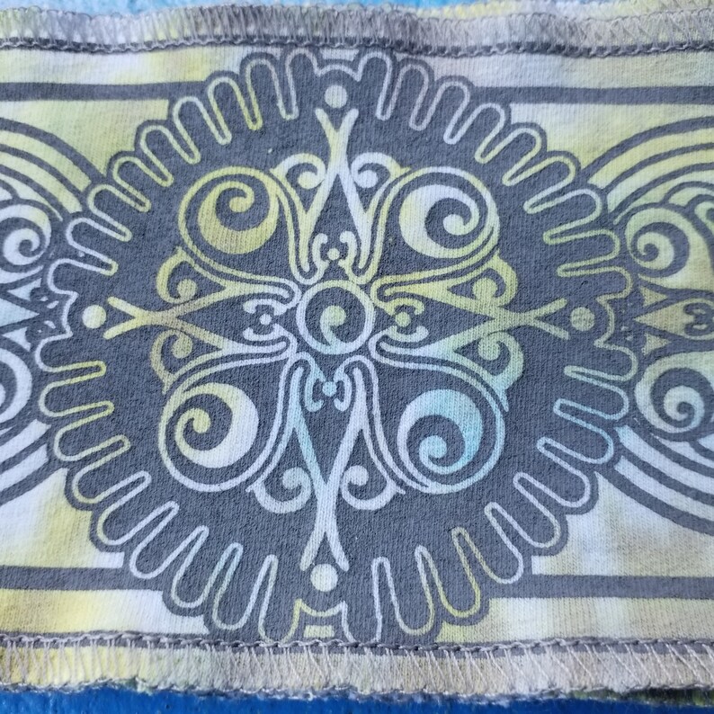 Op de afbeelding: Een grijze stof met een wit en geel mandala-ontwerp. De mandala heeft een cirkelvormig patroon met swirls en lijnen.