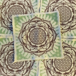 May include: Square fabric coasters with a brown and green mandala design. The coasters feature a geometric flower of life pattern in the centre and a decorative border. The background has a light green and white pattern.