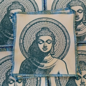 Peut inclure: Une pile de carrés de tissu blanc avec un motif brodé bleu d'un Bouddha en méditation. Chaque carré a un motif de mandala circulaire autour du Bouddha.