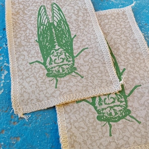 May include: Two beige fabric coasters with a green cicada insect design. The coasters have a patterned background and a white stitched border. The cicada illustration is detailed and centered on each coaster.