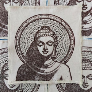 May include: Square, off-white fabric patch featuring a detailed brown illustration of Buddha with a halo. The image is centered and surrounded by a decorative border. The patch has a stitched edge.