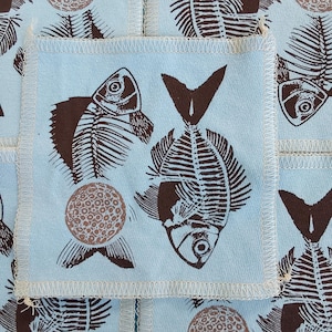 May include: A light blue fabric square with a dark brown screen-printed pattern featuring fish skeletons and circular dotted shapes. The square has visible off-white serged edges and is part of a larger repeating textile design.