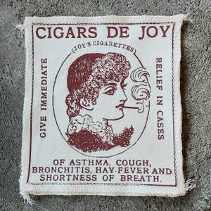 Può includere: Una toppa di tessuto color crema con un'illustrazione e un testo color amaranto. L'illustrazione raffigura una donna che fuma una sigaretta. Il testo include "CIGARS DE JOY" e "RELIEF IN CASES". La toppa è bordata da un bordo sfilacciato e il testo menziona il sollievo per asma, tosse, bronchite, febbre da fieno e mancanza di respiro.