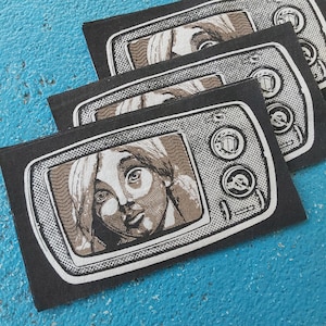May include: Several black fabric patches with a retro television design. Each patch shows a sepia-toned portrait inside the TV screen, with control knobs and a speaker grill. The patches are laid out on a blue surface.