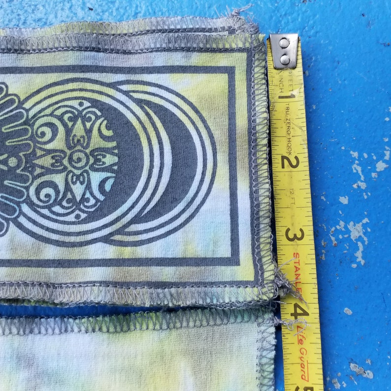 Op de afbeelding: Een close-up van een tie-dye stof met een grijs en wit ontwerp. De stof wordt gemeten met een meetlint, wat een lengte van 10 cm laat zien.