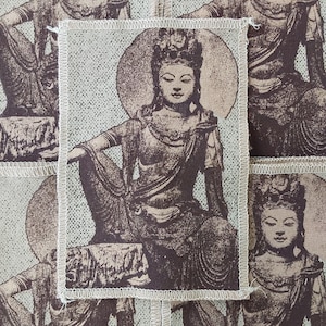 May include: A rectangular fabric patch featuring a dark brown screen-printed illustration of a seated deity with a circular halo against a light beige background. The patch has a visible white overlock stitched edge on all four sides.