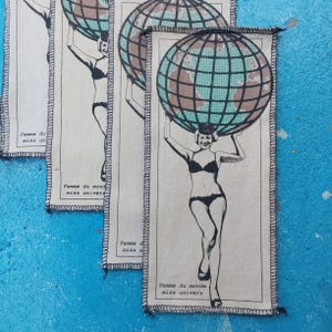 May include: Several rectangular fabric patches with a black and white illustration of a woman in a bikini holding a globe above her head. The text "Femme du monde miss univers" is printed on the patches.