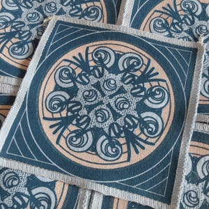 Puede incluir: Parches cuadrados de tela con un diseño circular repetitivo. El diseño presenta un centro de color melocotón claro con un patrón arremolinado, rodeado por un borde azul oscuro. Los bordes están acabados con un ribete plateado.