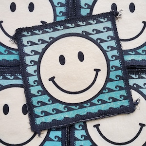 Pode incluir: Um remendo de tecido azul e branco com um rosto sorridente no centro. O remendo tem uma borda preta e é costurado ao redor das bordas.