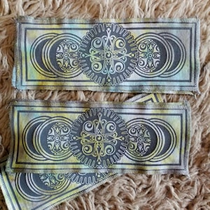 May include: Two fabric bookmarks with a repeating pattern of a stylized moon and sun design. The bookmarks are in shades of green, yellow, and black.