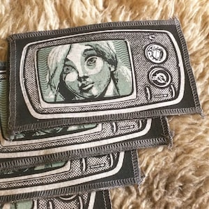 Peut inclure: Un ensemble de patchs en tissu noir et blanc avec un design de télévision vintage. L'écran de télévision montre un gros plan du visage d'une femme.