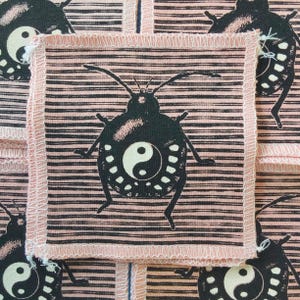 May include: Square fabric patches with a black insect design featuring a yin-yang symbol on a pink and black striped background. The patches have a pink border with a frayed edge. The design is repeated across the fabric.