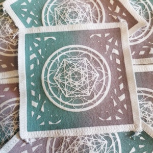 May include: A square patch with a white geometric design on a teal and brown gradient background. The patch has a white border with a frayed edge.