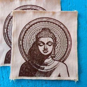May include: A square fabric patch featuring a dark brown screen-printed illustration of a Buddha figure with closed eyes, centred within a detailed circular mandala pattern on a light beige background with finished stitched edges.