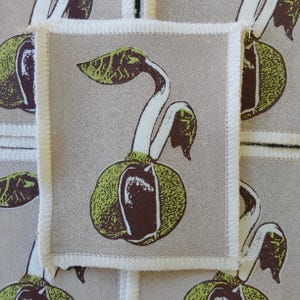 May include: A rectangular fabric patch with a beige background featuring a screen-printed graphic of a sprouting seed in shades of dark brown and lime green. The patch has a white stitched border and is layered over a matching patterned fabric.