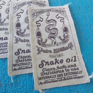 Peut inclure: Plusieurs écussons rectangulaires en tissu avec un design vintage. Les écussons présentent une illustration de serpent, le texte "Snake Oil" et d'autres textes. Les écussons ont un fond crème avec du texte et des illustrations marron foncé.