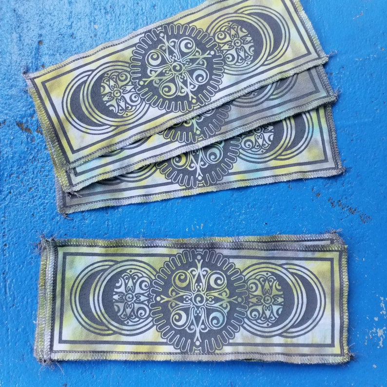 Op de afbeelding: Een set van vier stoffen bladwijzers met een herhalend patroon van een mandala-ontwerp in tinten groen, geel en zwart. De bladwijzers zijn gestikt met een zigzagsteek langs de randen.