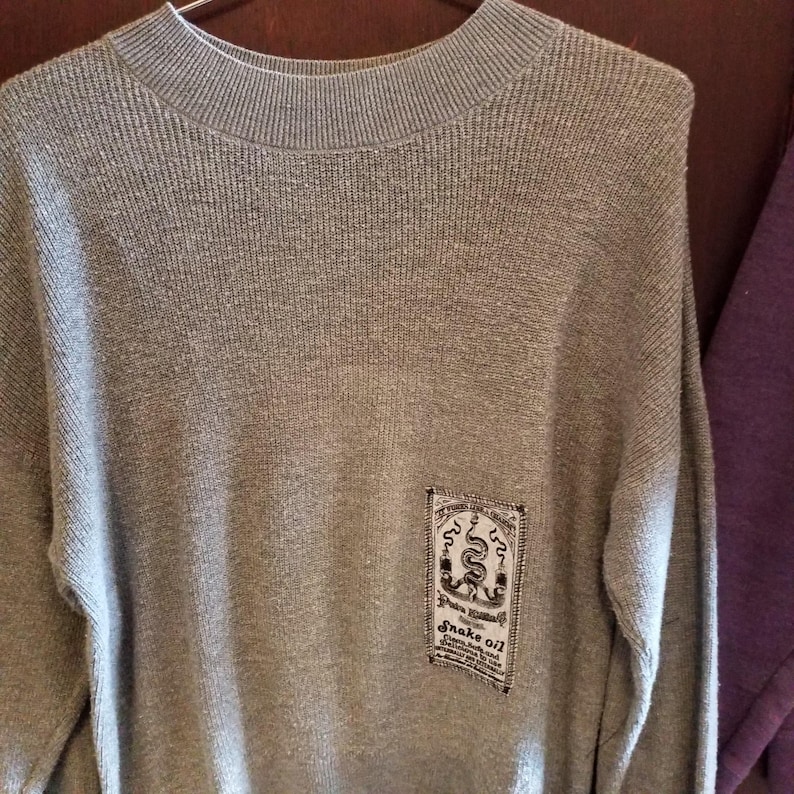 Op de afbeelding: Een grijze trui met lange mouwen met een klein rechthoekig patch aan de linkerkant van de borst. De patch heeft een zwart-witte afbeelding van een slang en de tekst "Snake Oil" erop gedrukt.